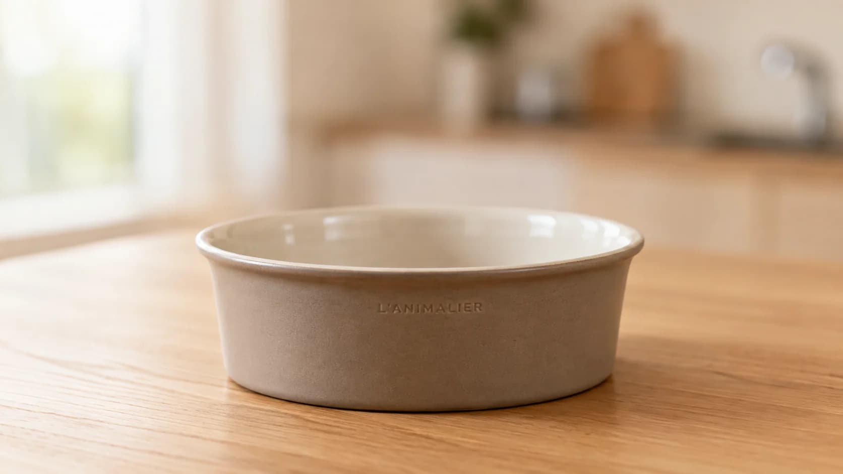 Bol de gres taupe con interior esmaltado crema y el nombre de la casa grabado en el barro, sobre una mesada de roble claro con la cocina desenfocada al fondo.