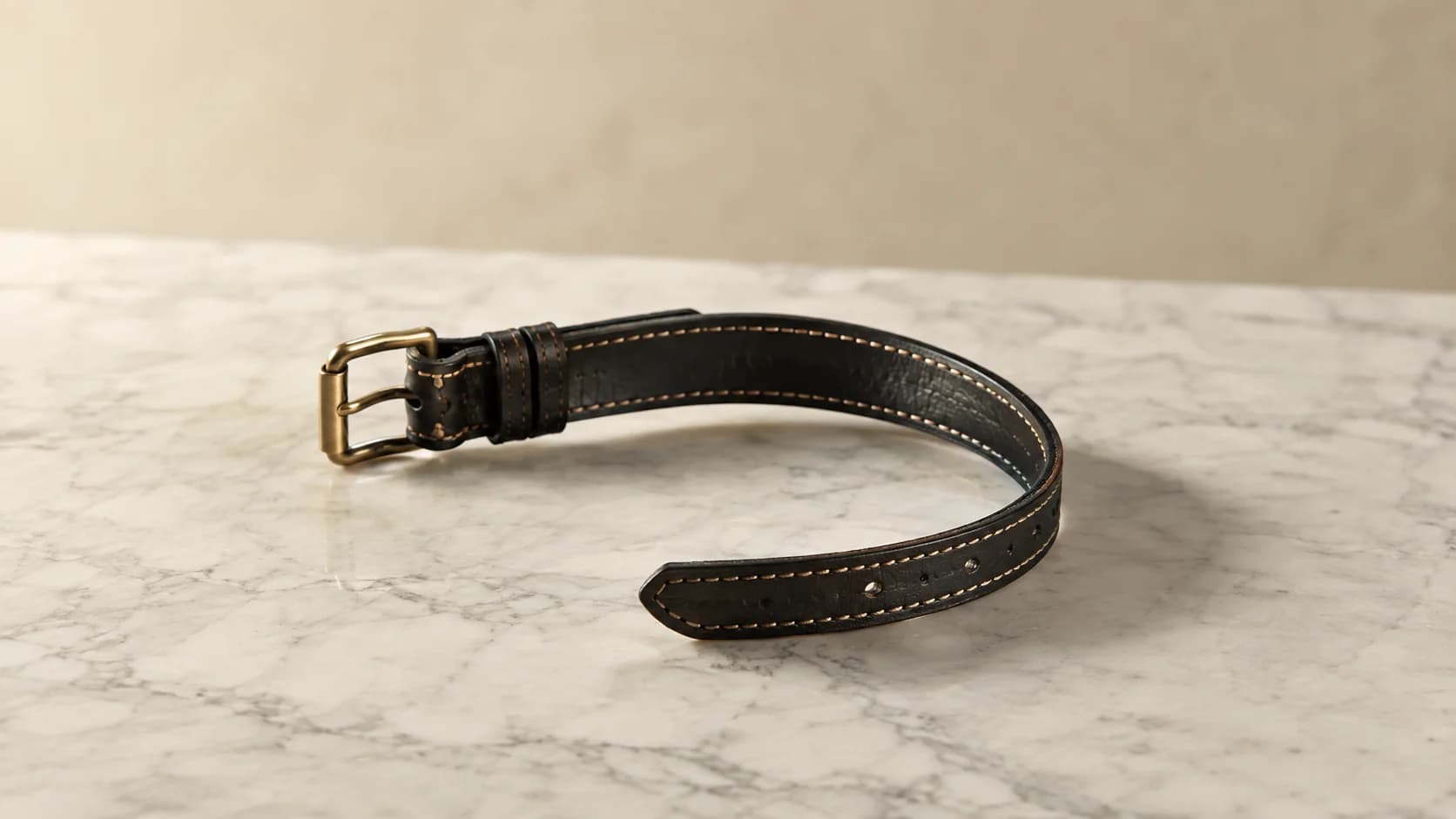 Collar de cuero de guarnicionería negro con hebilla de latón macizo, abierto sobre mármol pulido.