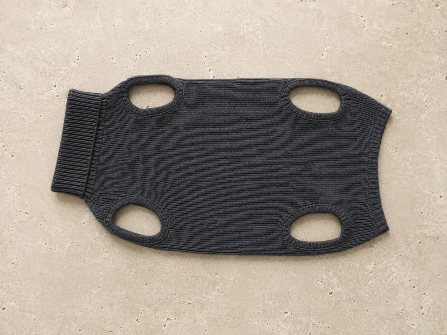 Suéter de cachemira gris carbón para perro, extendido en plano sobre travertino crema, con sus cuatro aberturas de patas a la vista.