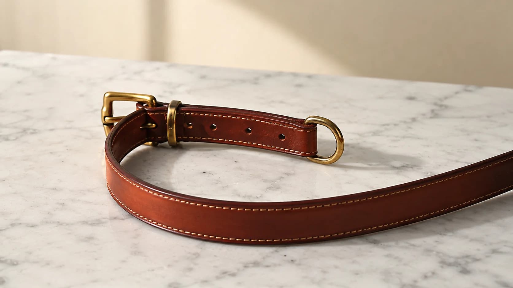 Collar de cuero coñac flor entera con hebilla de latón macizo y costura de silla, en curva abierta sobre una superficie de mármol de Carrara.