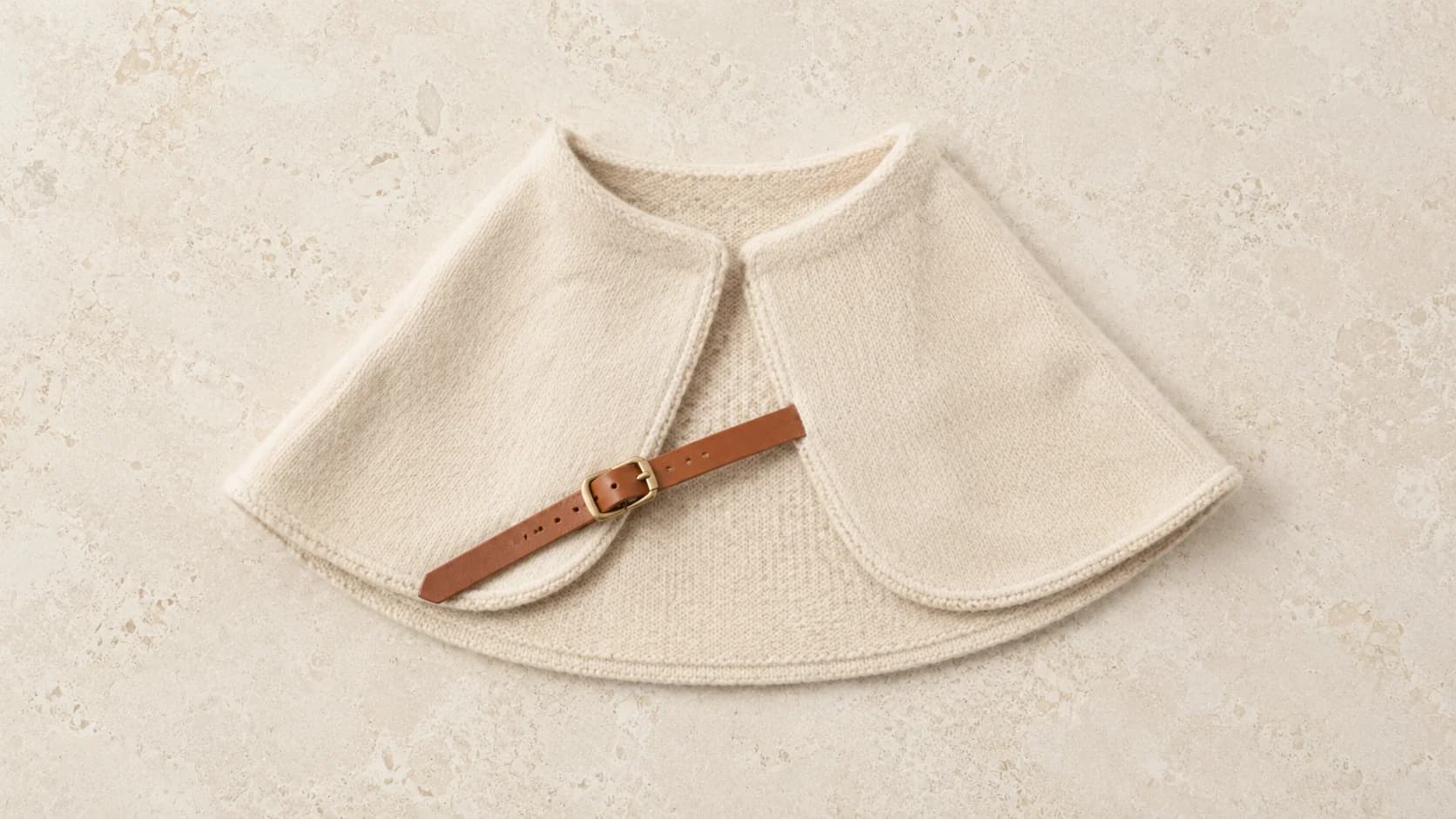 Capa de baby alpaca crema para perro, extendida en abanico sobre piedra caliza pálida, con la tira de cuero coñac cruzando en diagonal.