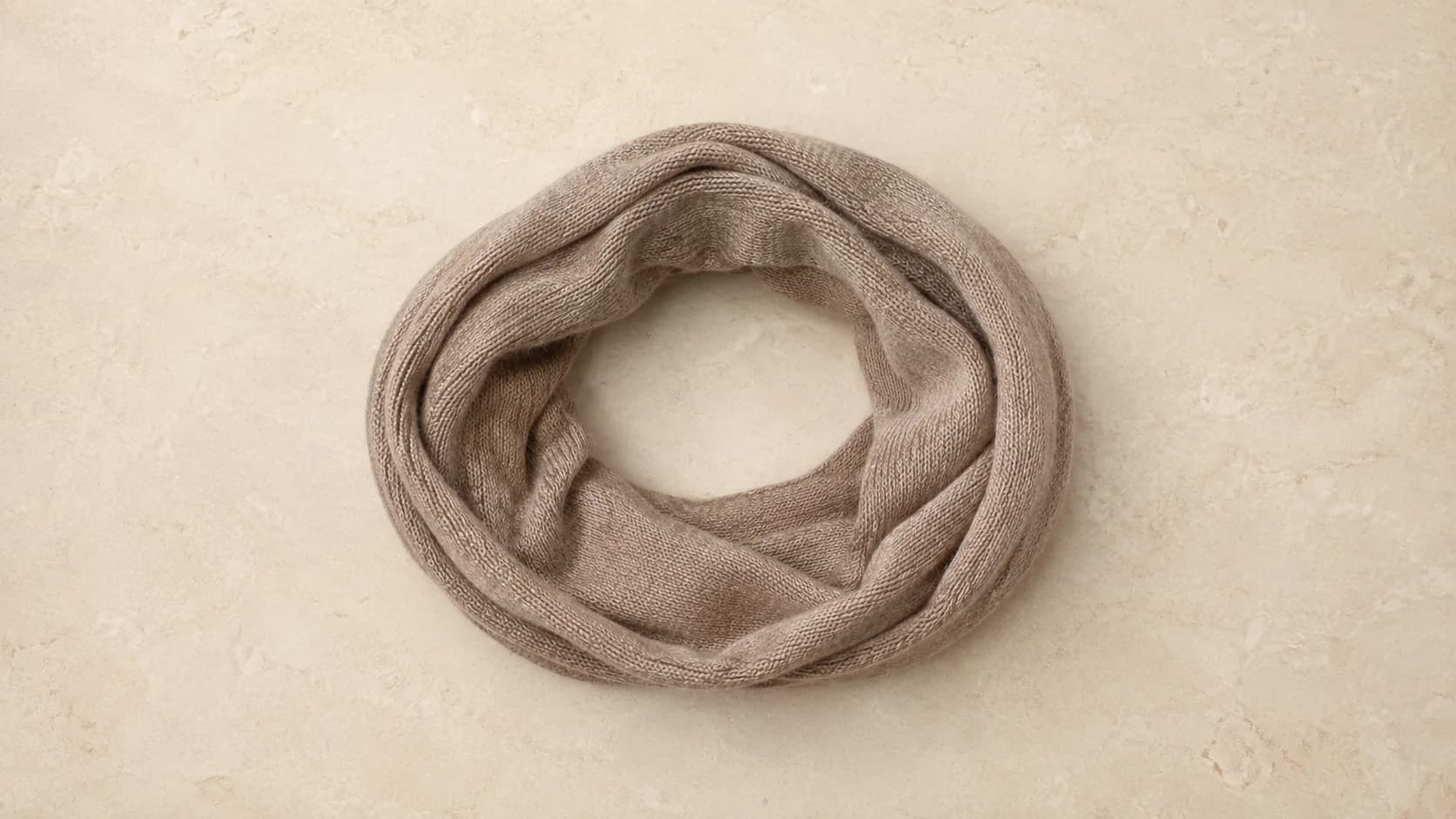 Cuello de cachemira taupe sin costuras, apoyado como un aro abierto sobre piedra clara.