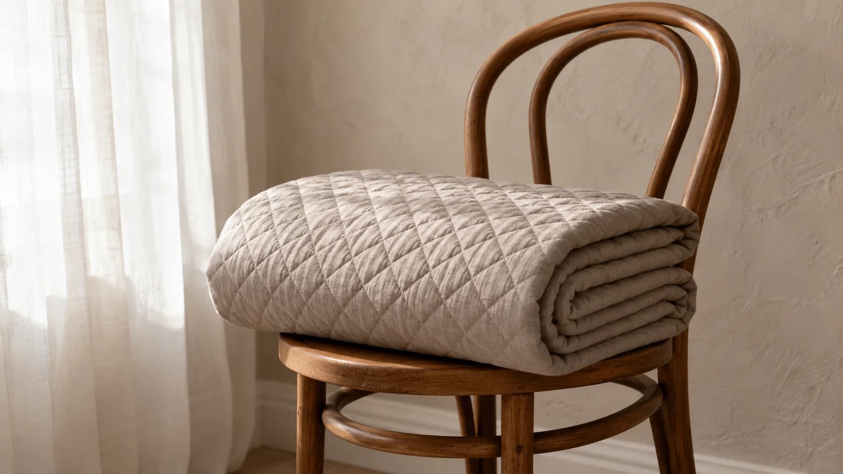 Cubrecama de lino matelasseado en taupe, doblado en tercios sobre el asiento de una silla de madera curvada, con la luz de invierno marcando cada rombo.