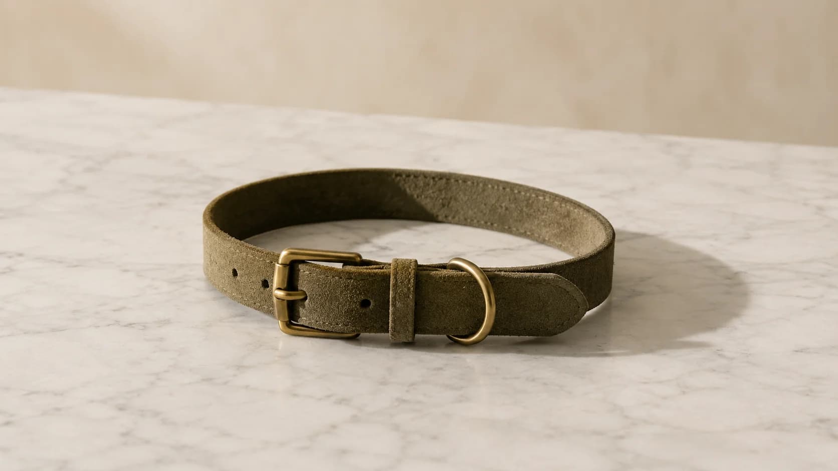 Collar de ante italiano oliva extendido sobre mármol, con herrajes de latón mate.